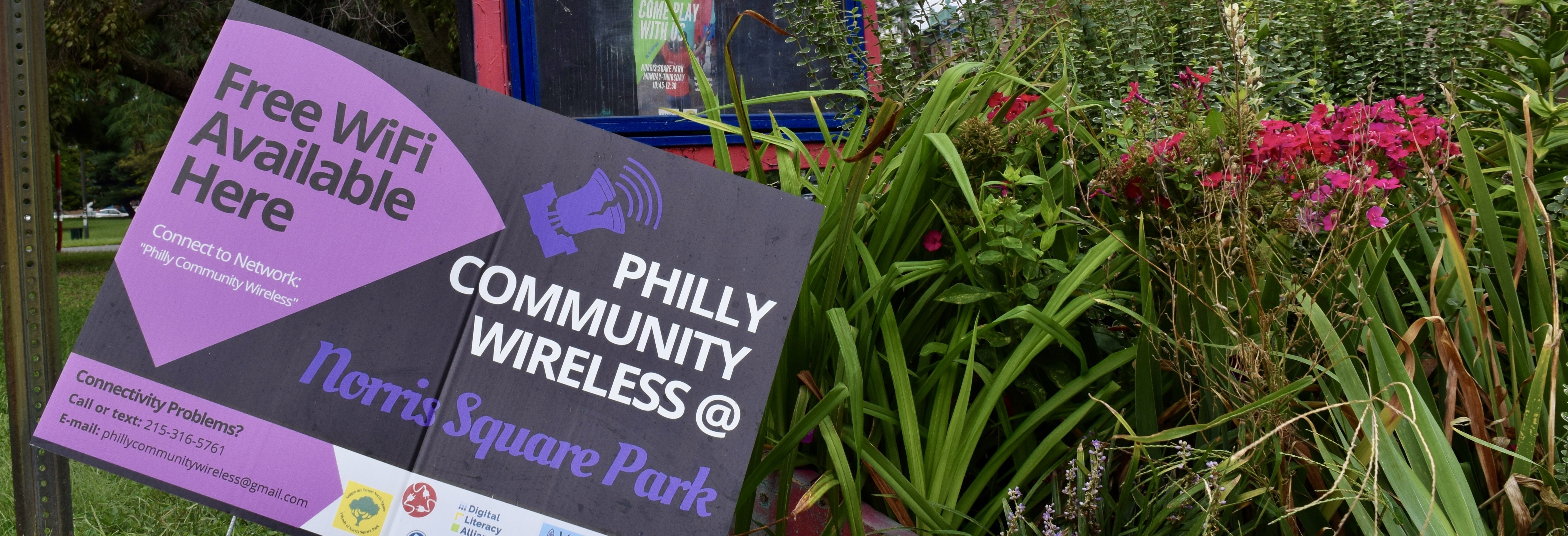 A Philly Community Wireless sign amongst some flowers in Norris Square Park