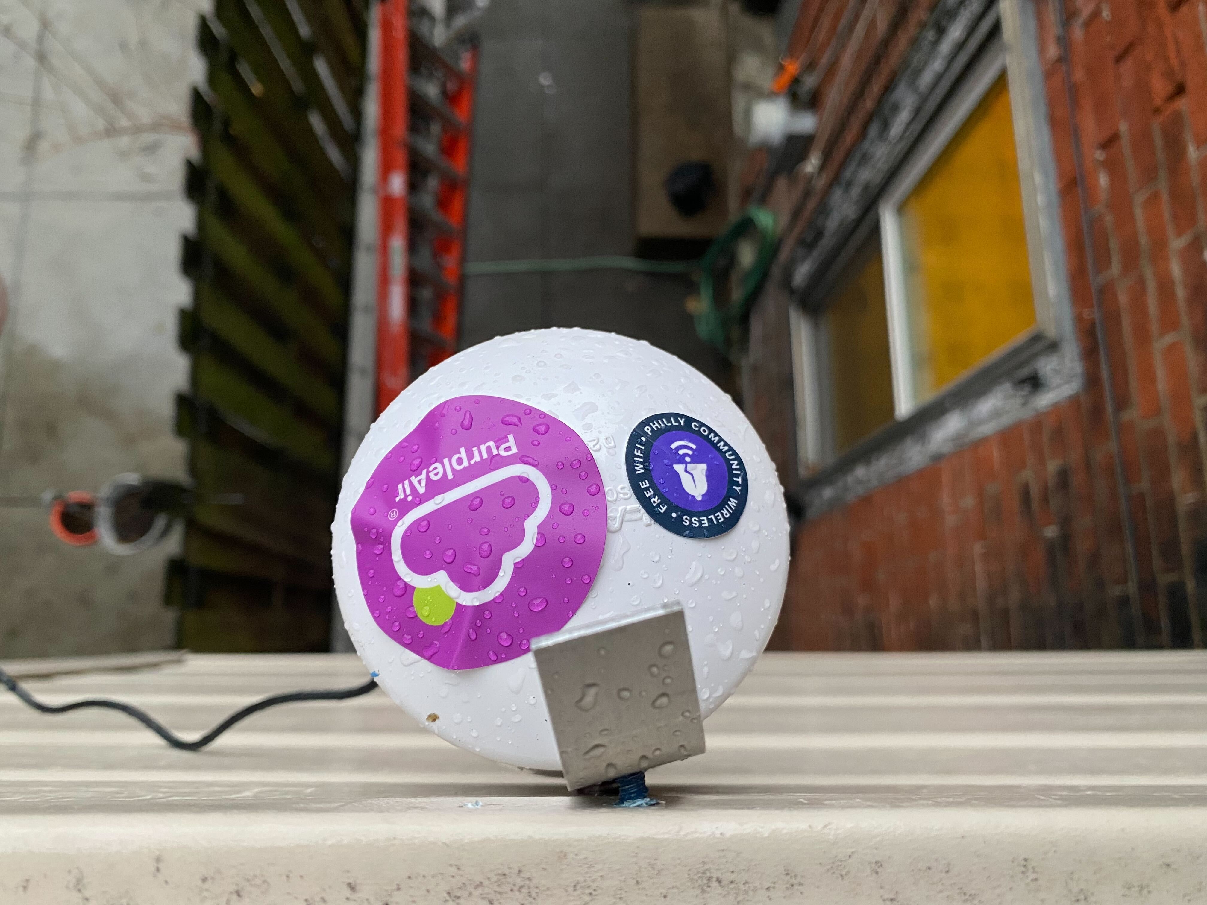 An ariel view of a white circular device with stickers saying Purple Air and Philly Community Wireless attached to a house