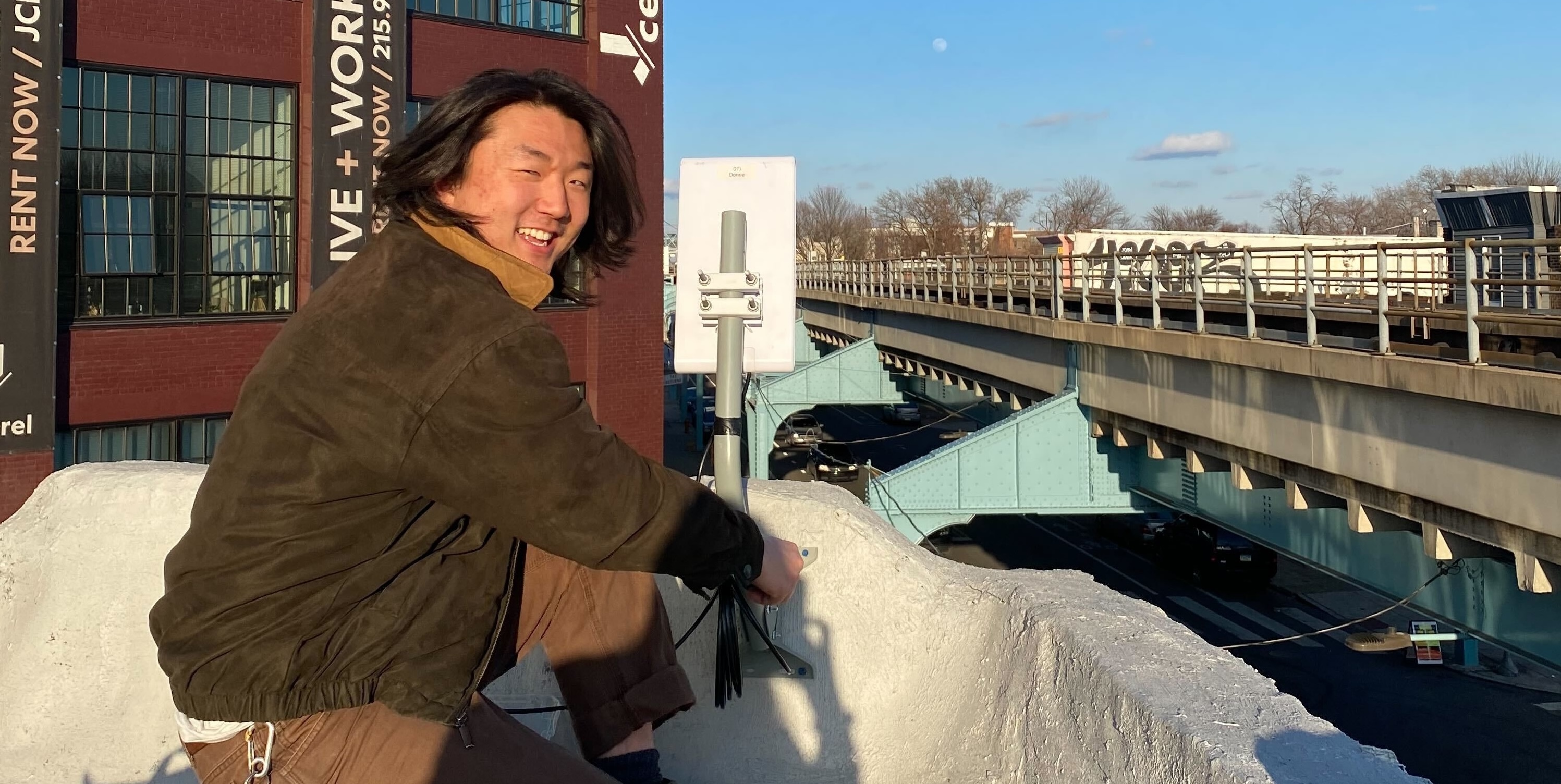 A man on a roof smiling while he secures a pole mount with an access point device pointing towards the street and subway tracks.