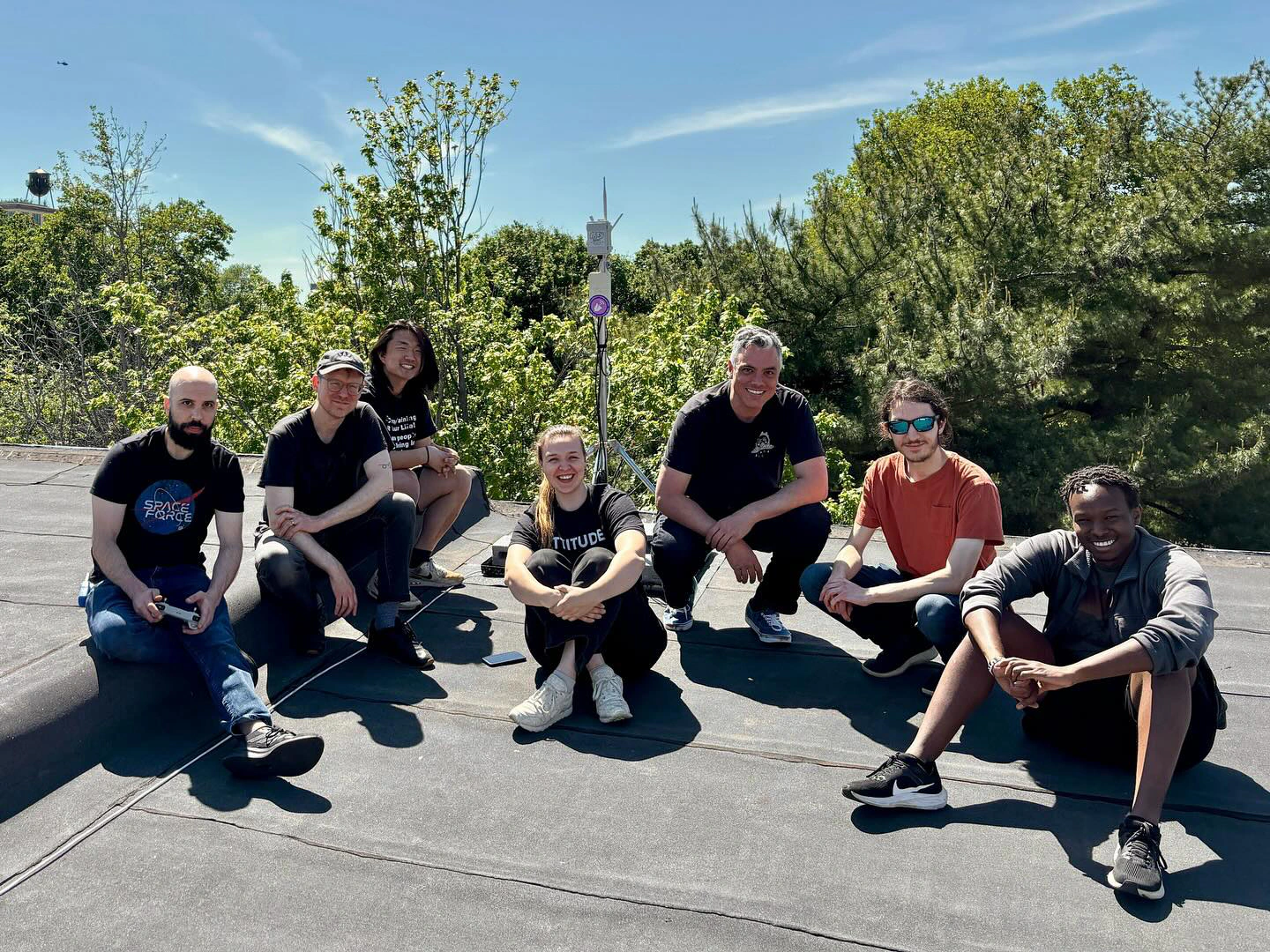 7 people posing for a group photo on a roof with a device and tall trees behind them.