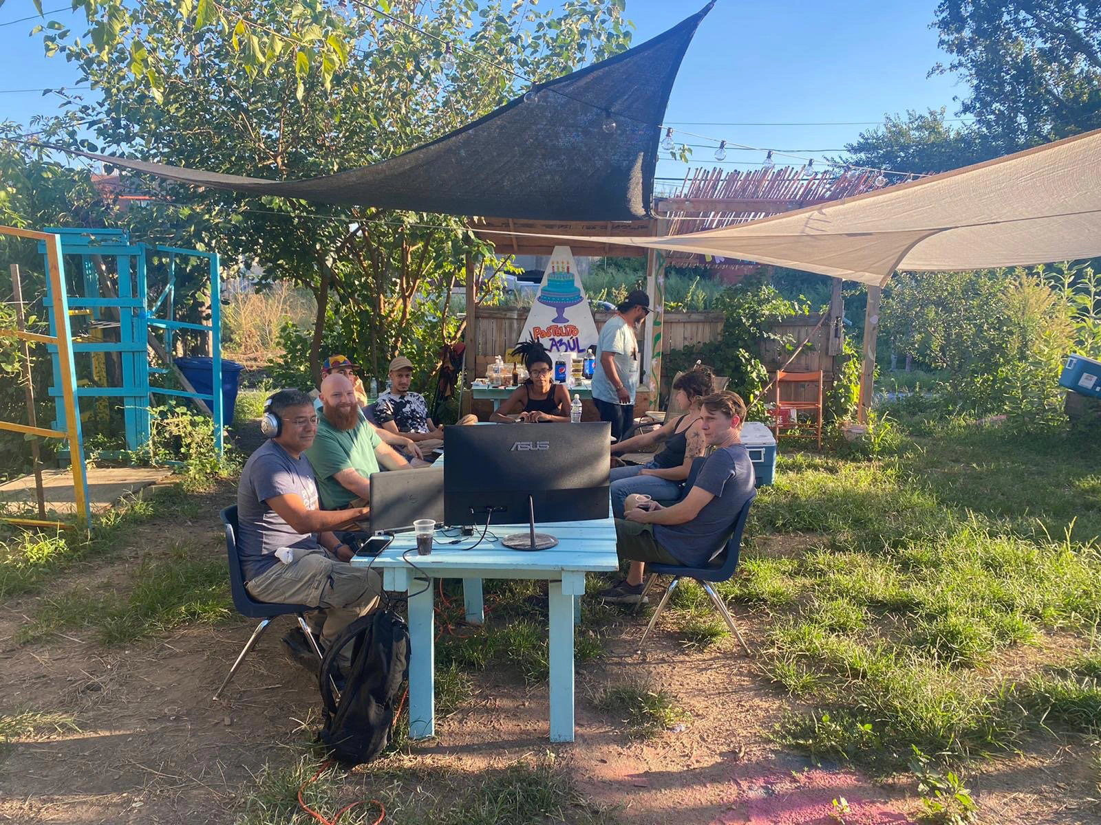 Vecinos sentados alrededor de mesas bajo un toldo en un jardín comunitario.