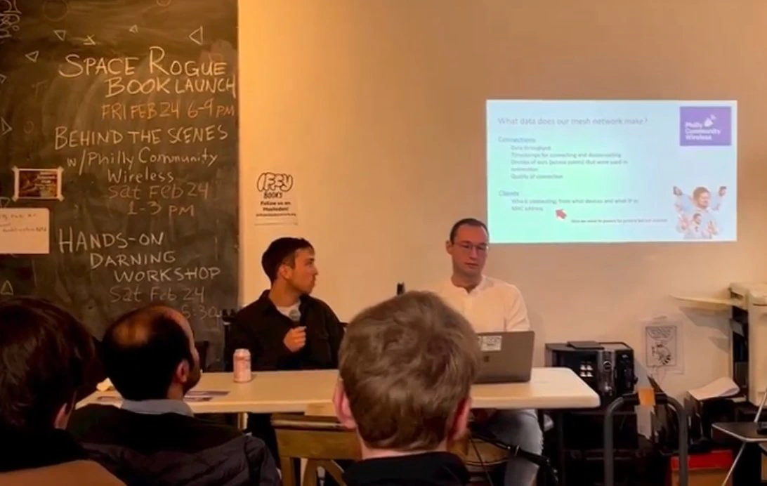 Dos presentadores frente a una computadora portátil, junto a una pizarra y una diapositiva proyectada, en Iffy Books.