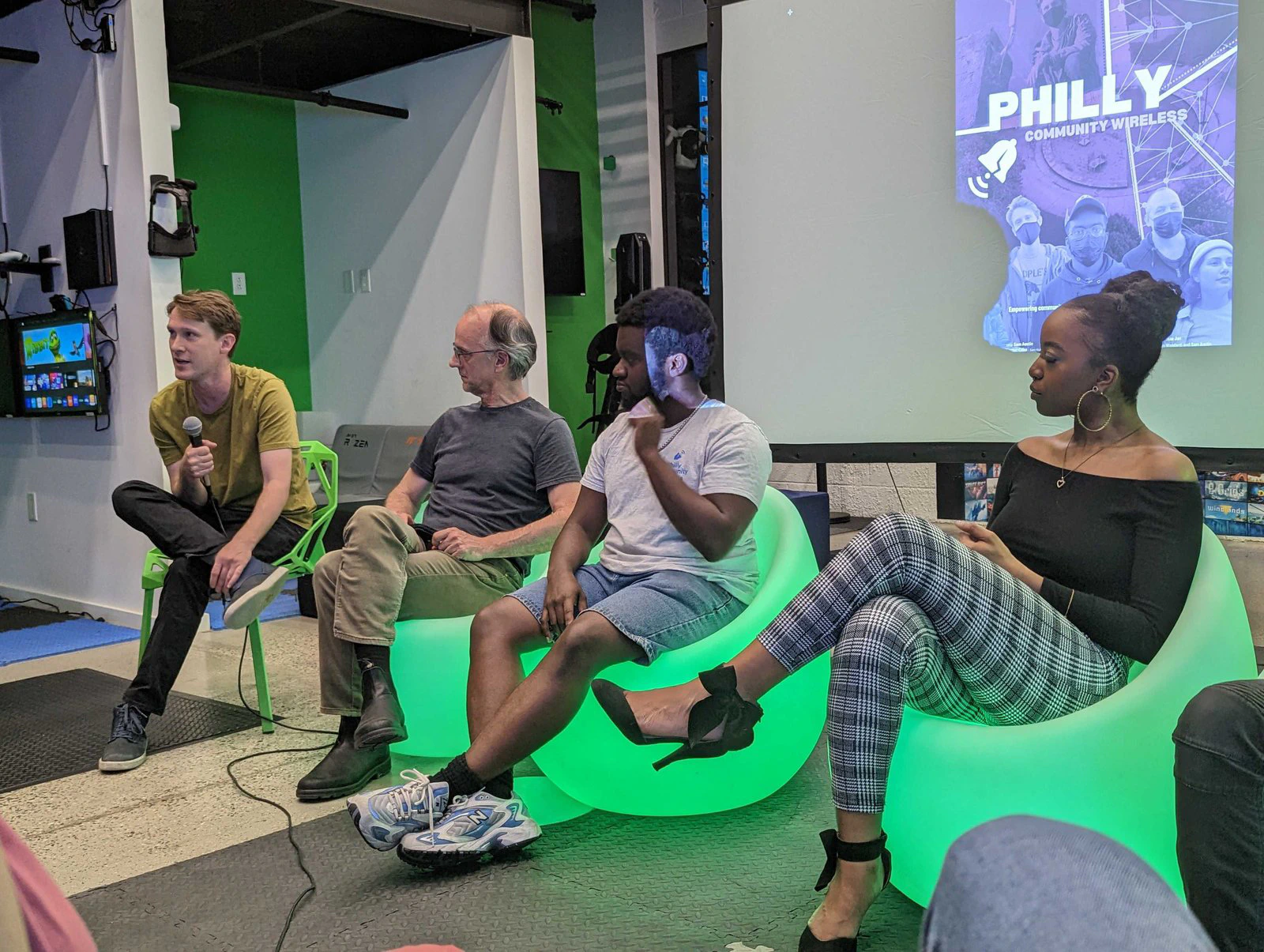 Cuatro panelistas sentados en sillas verdes en un estudio, con una diapositiva de Philly Community Wireless detrás.
