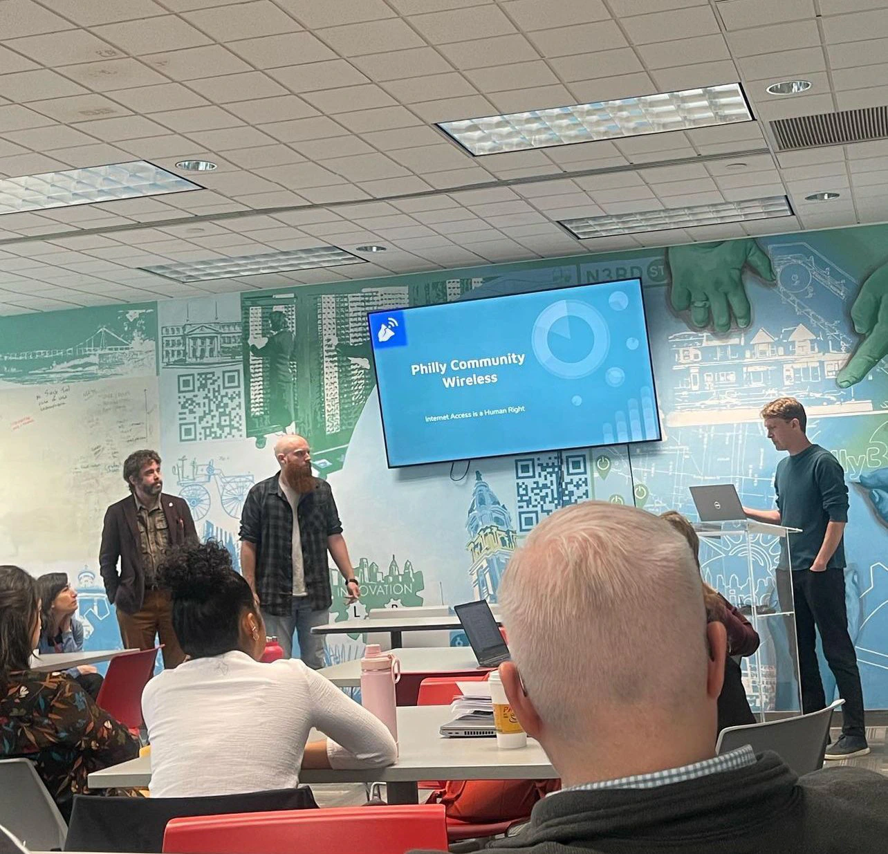 Un presentador habla junto a una diapositiva proyectada de Philly Community Wireless en una sala de reuniones.