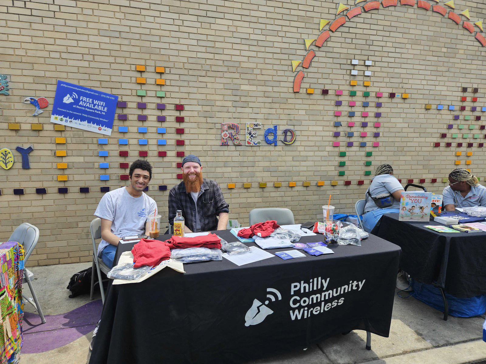 Dos voluntarios en una mesa de Philly Community Wireless con una pancarta de Wi-Fi gratis, frente a una pared de mosaico.