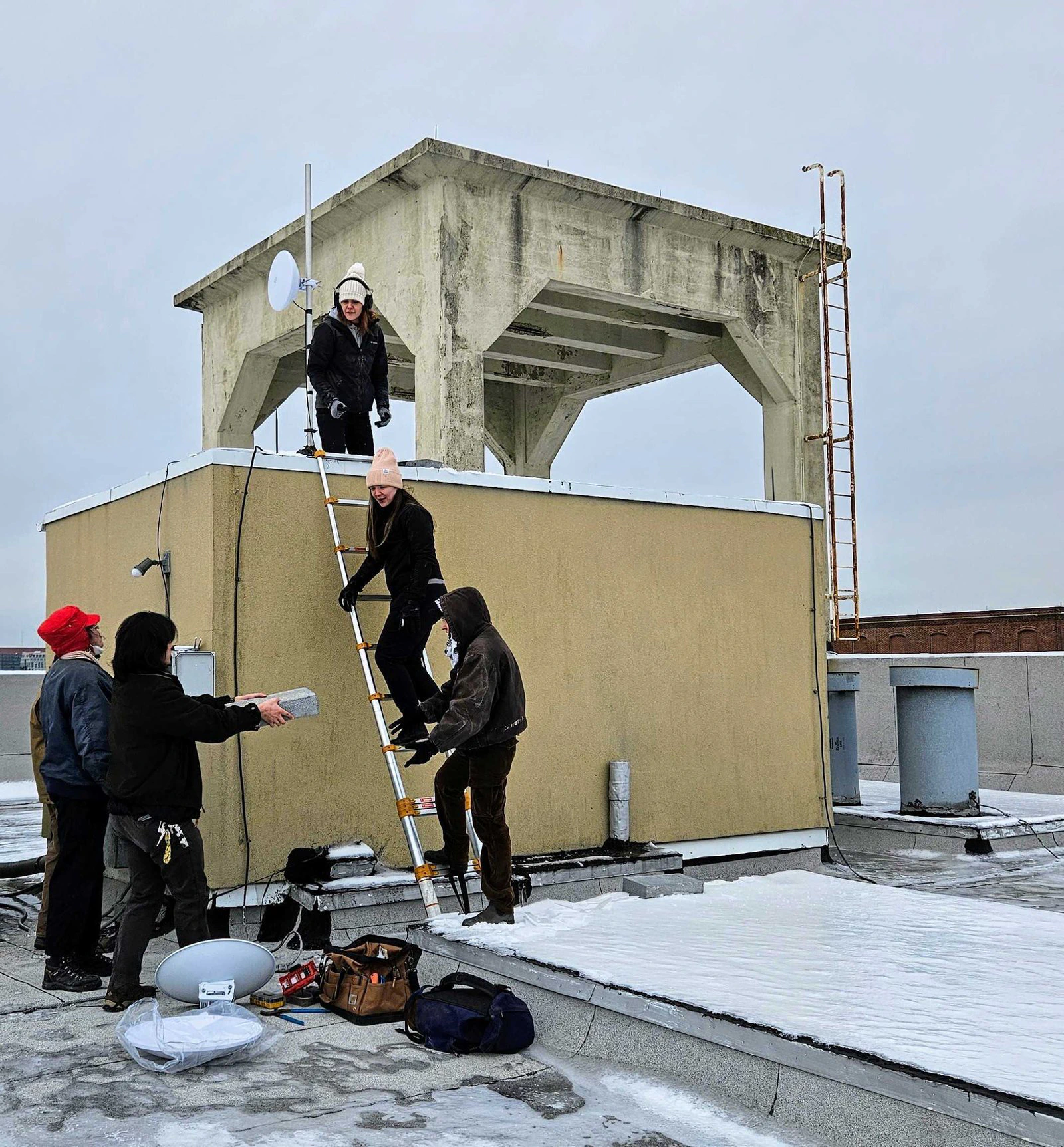 Técnicos sobre una escalera de mano y en la caseta de acceso de un techo instalan una antena en un edificio de HACE.