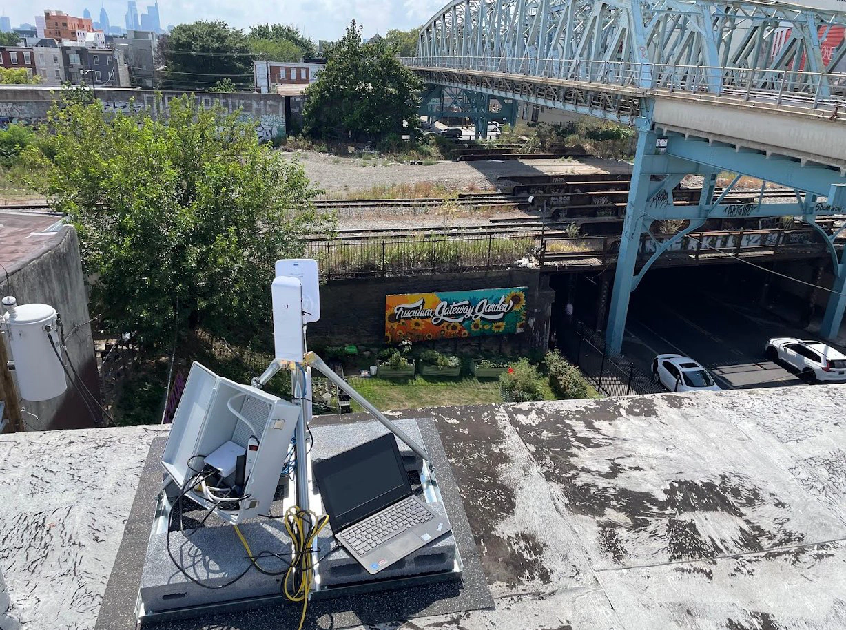 Una antena, una caja de equipos y una computadora portátil en un techo, con la línea Market-Frankford al fondo.