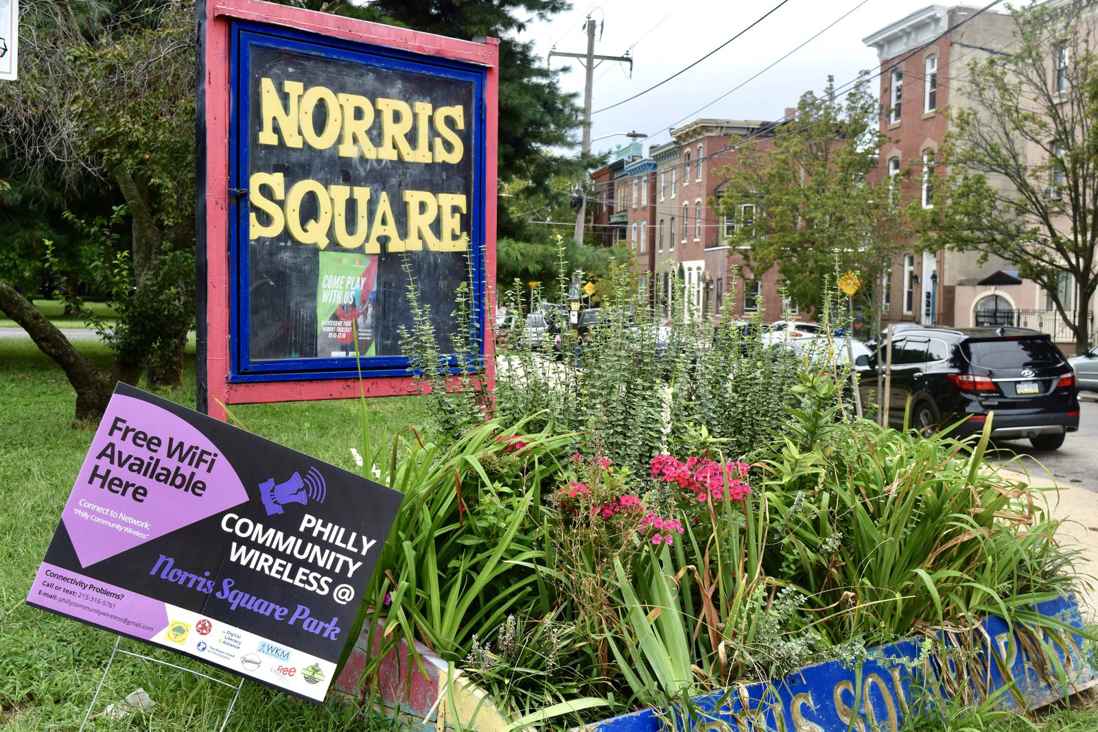 El letrero de Norris Square Park y, delante de él, un cartel de jardín de Philly Community Wireless que dice "Free WiFi Available Here" (WiFi gratis disponible aquí).