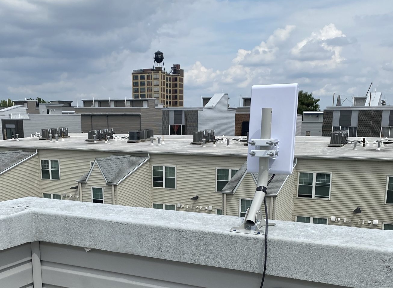 An access point device mounted on the edge of a roof pointing towards houses and a tall building in the background.