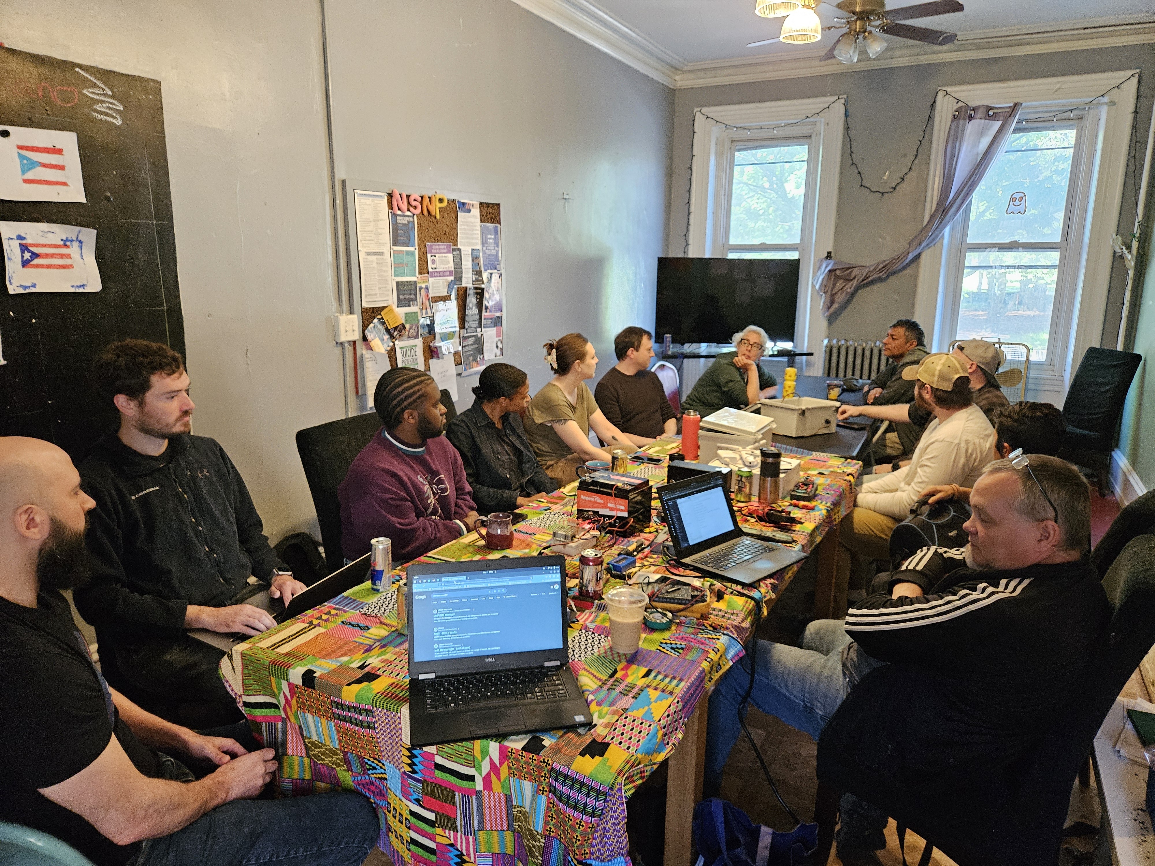 12 people sitting around a table indoors with laptops and tech equipment on the table.