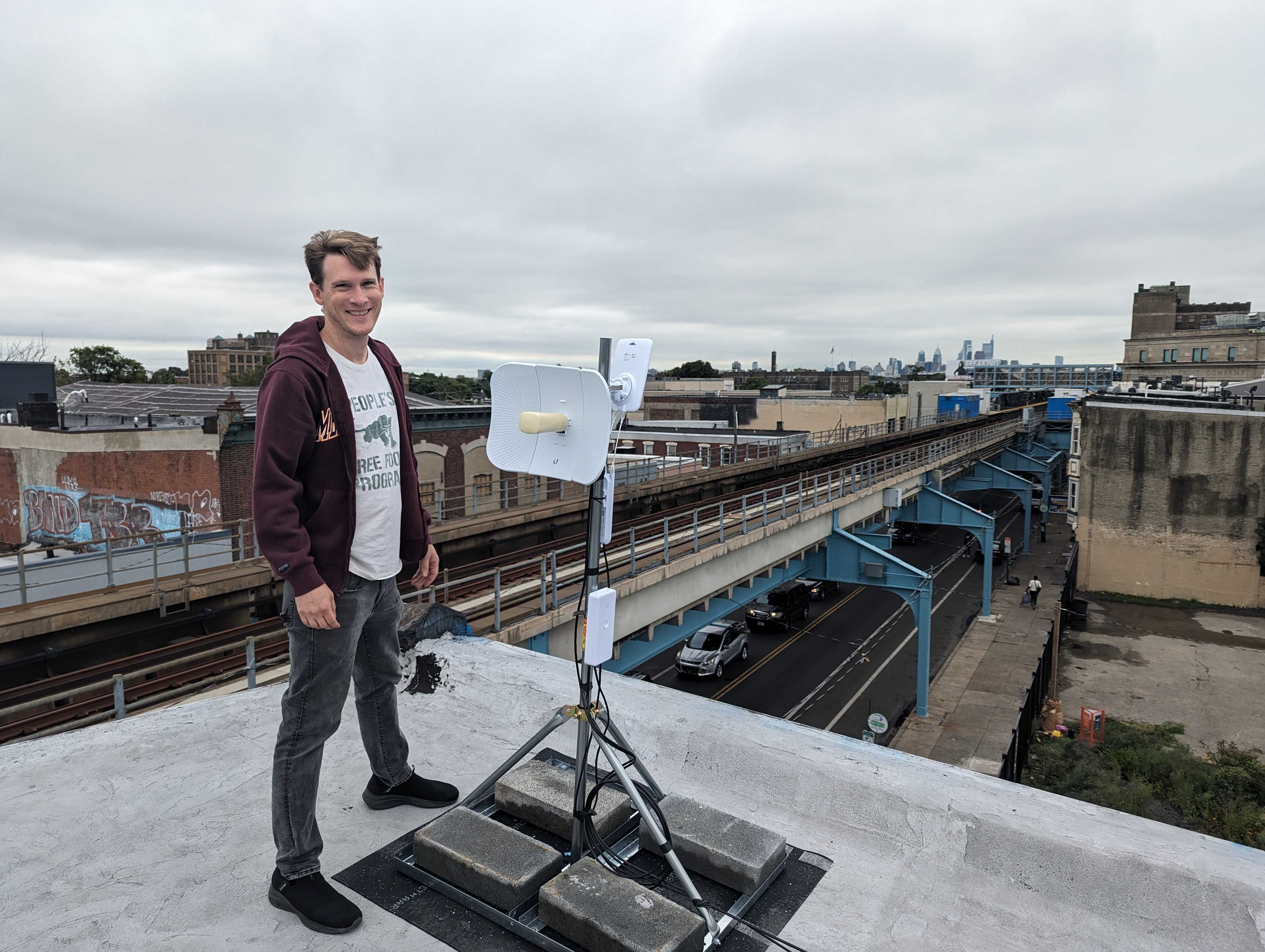 A man smiling standing next to a structure with a LiteBeam and access point device on a roof, with subway tracks and the street in view behind him.