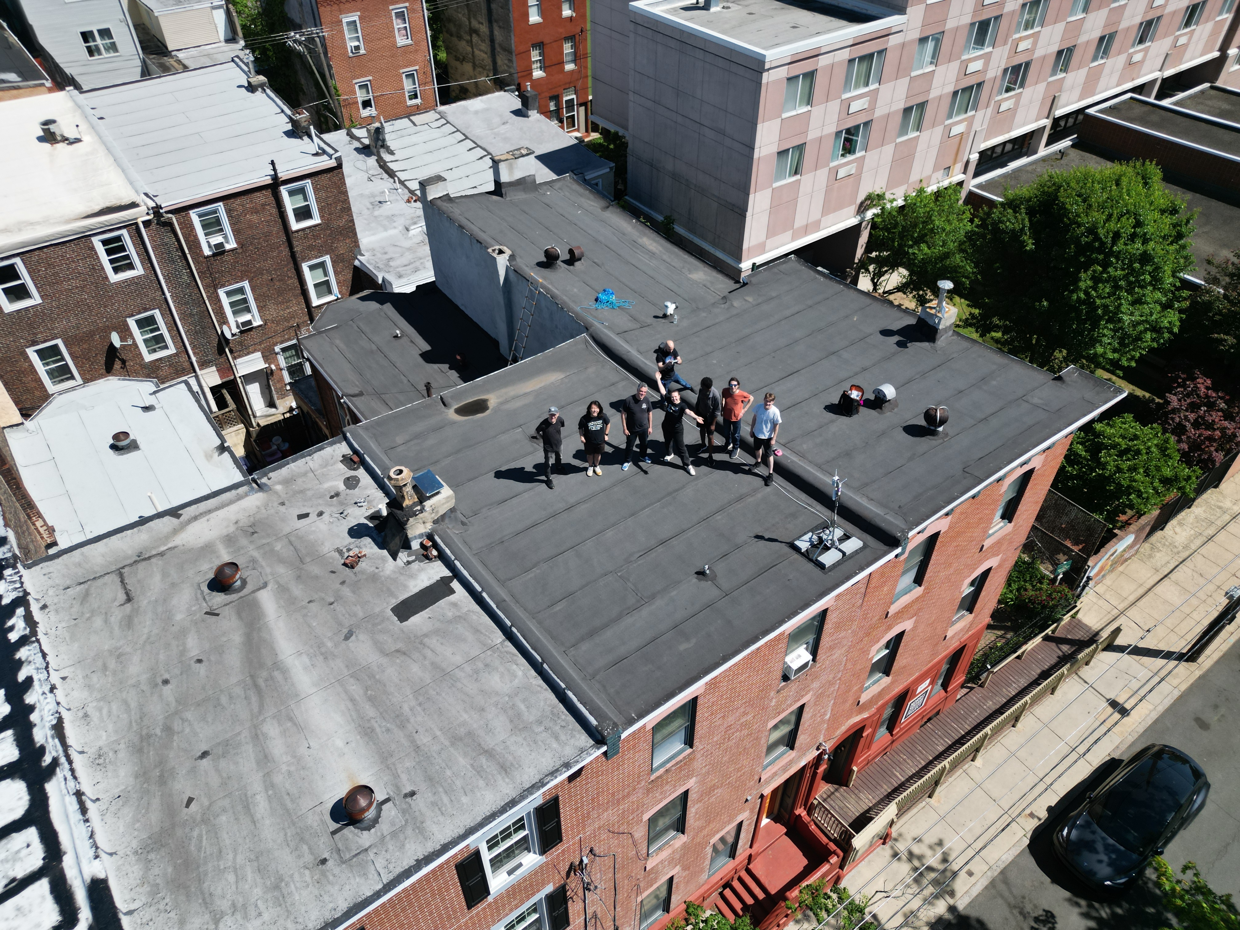 A drone shot of 8 people posing on a roof showing several building rooftops and the sidewalk.