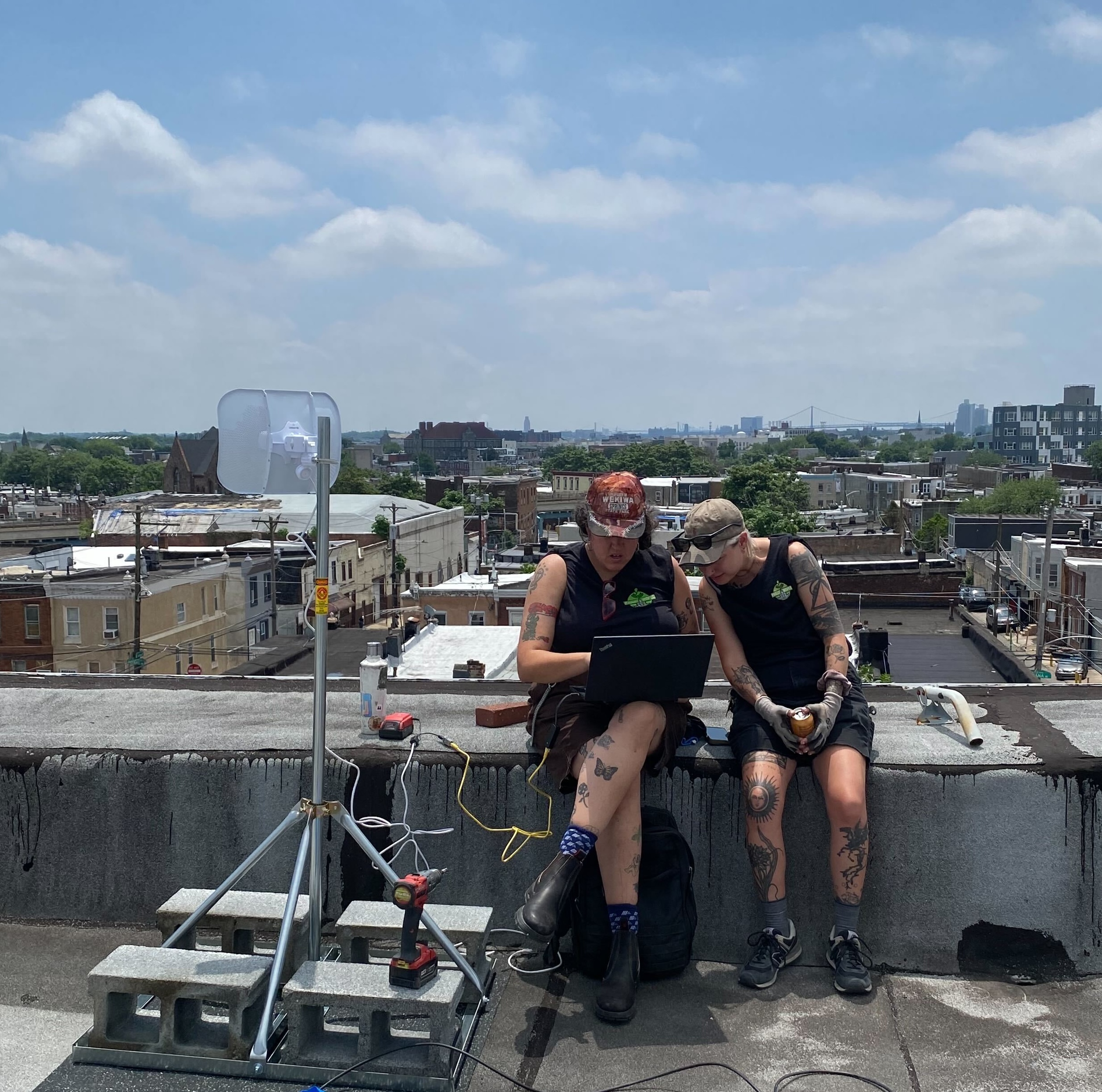 Two PhillyWisper staff sitting on a roof looking at a laptop, with a structure and LiteBeam device next to them.