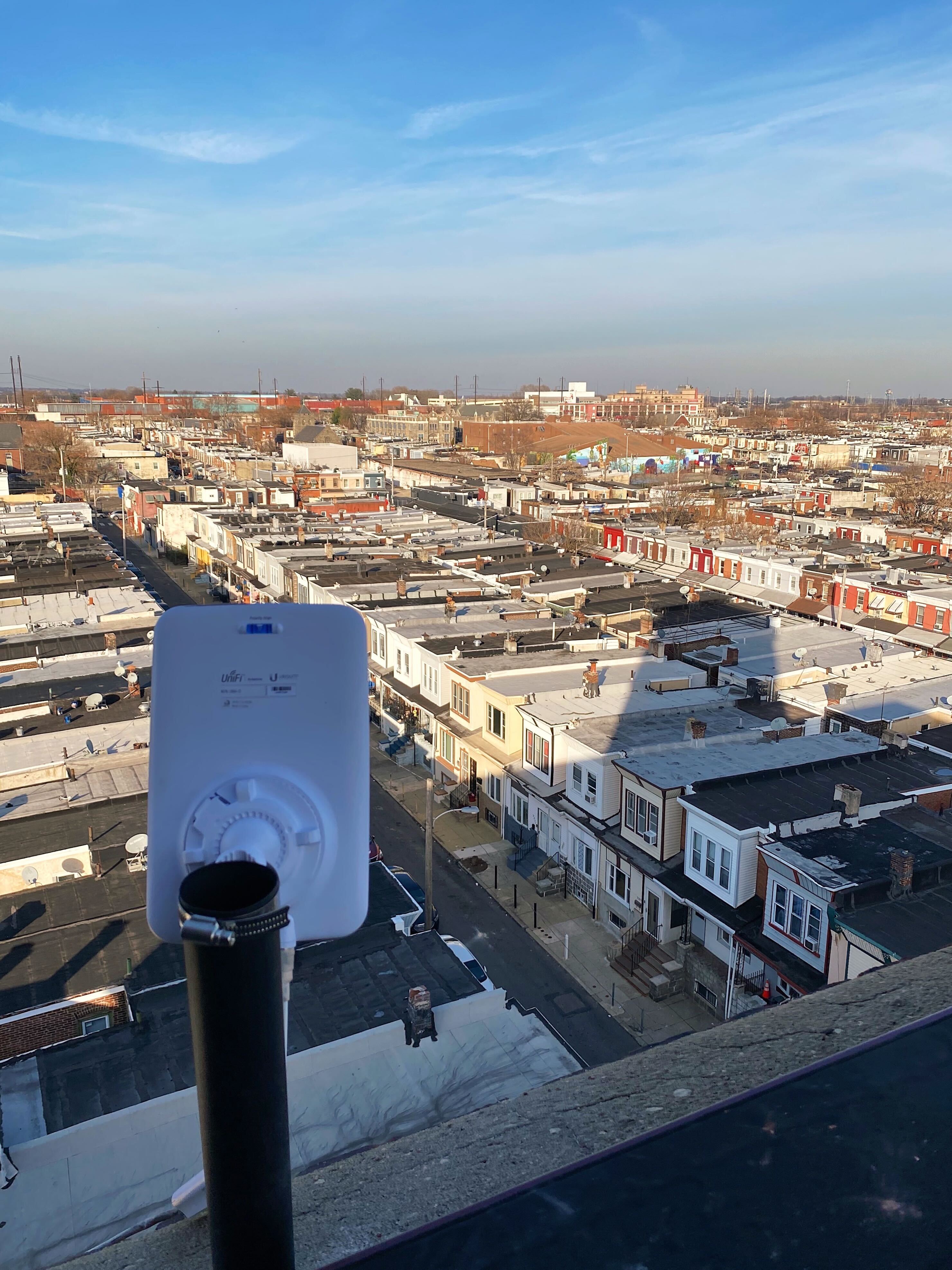 A white rectangular access point device pointing down from high up towards blocks of rowhomes.