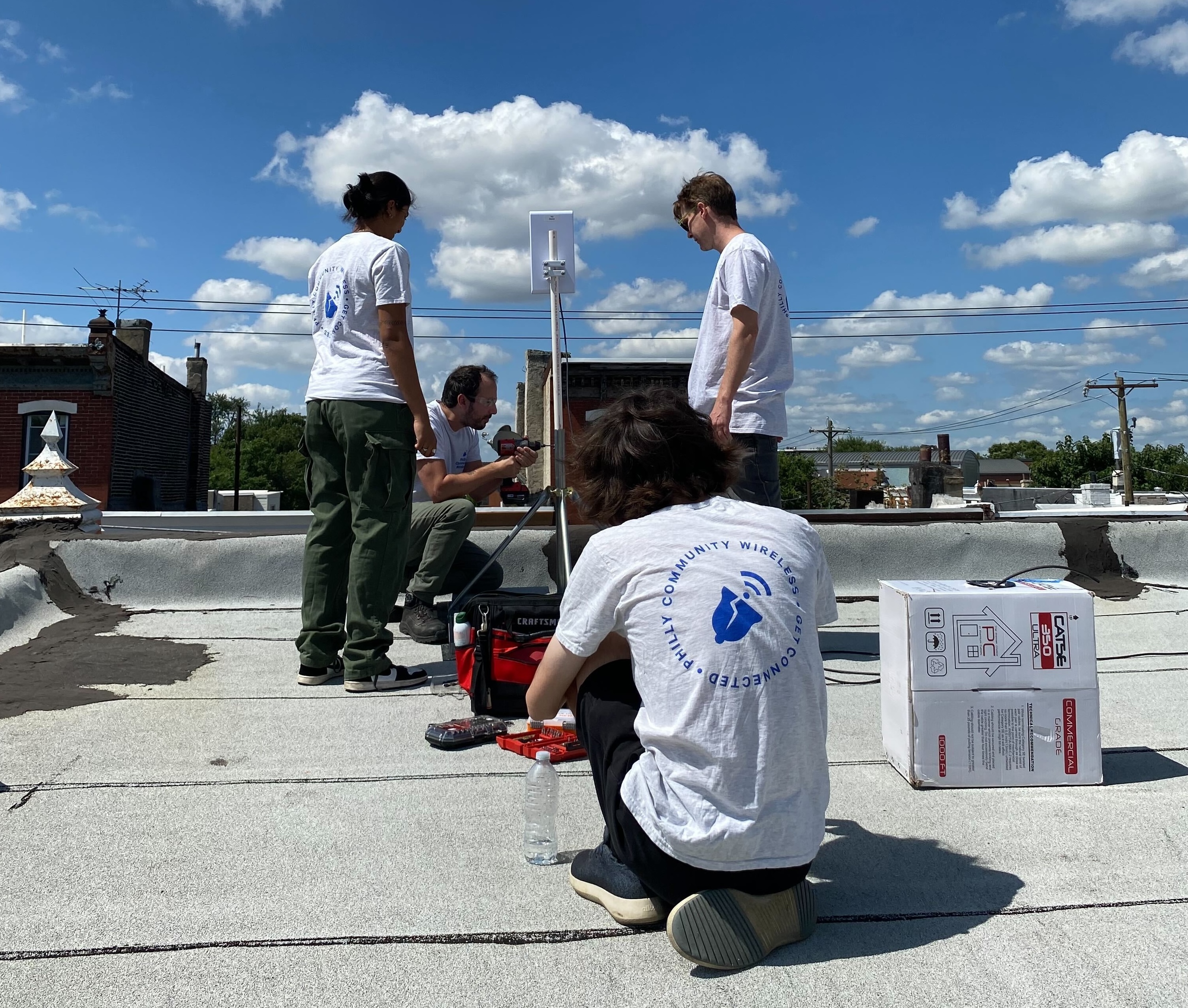 4 PCW Volunteers on a roof, with one using a drill to secure a pole that the access point device is on.
