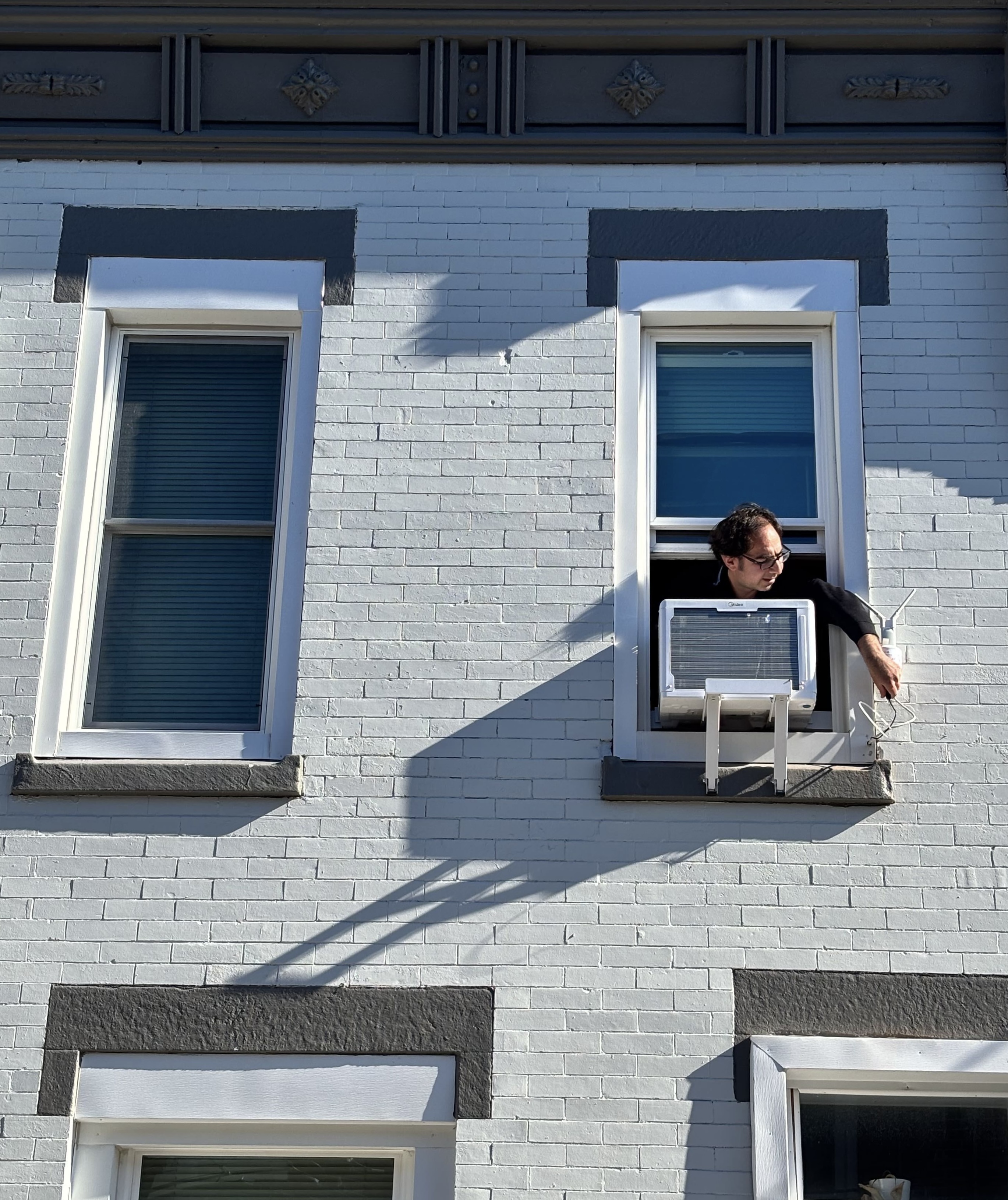 A man reaching out of a second floor window with one arm to adjust a device next to an air conditining unit.