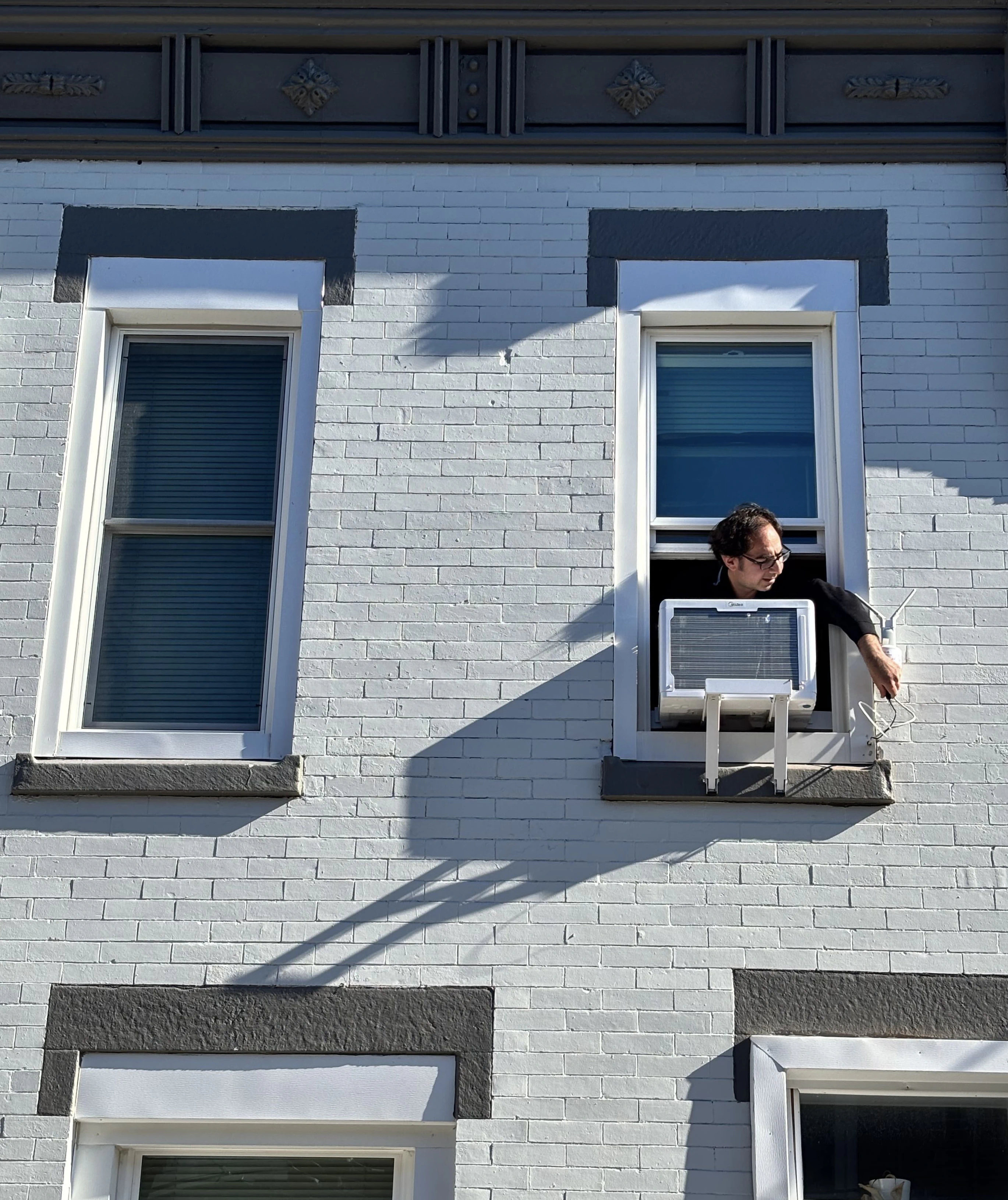 Un hombre asomado por la ventana del segundo piso con un brazo para ajustar un dispositivo junto a una unidad de aire acondicionado.