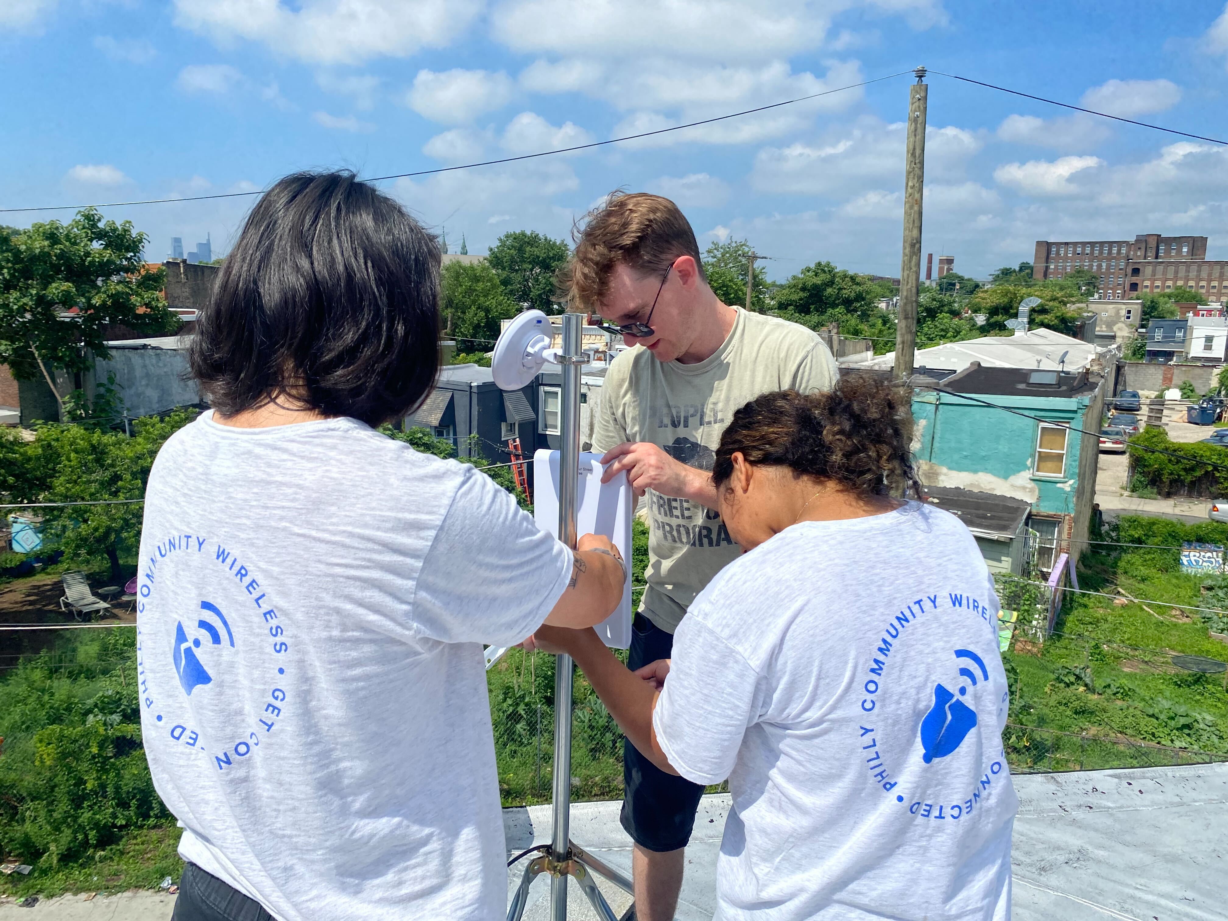 3 people on a roof securing an access point device to a pole with a garden in the background at street level.